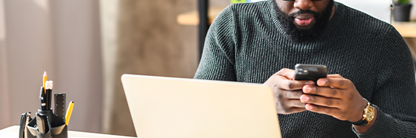 Man texting on a smartphone while sitting at a desk with his laptop open